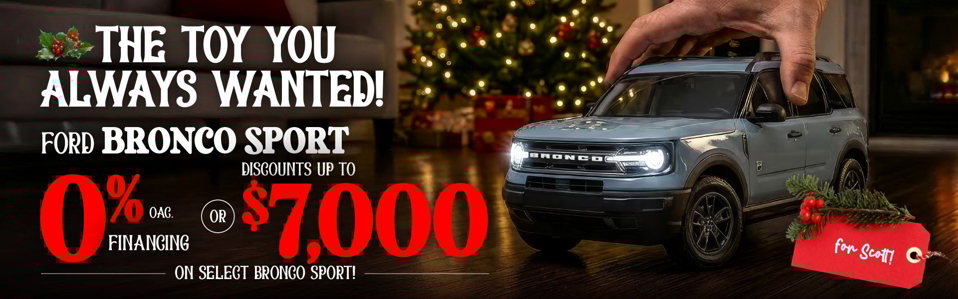 A hand lowers a toy-sized light blue Ford Bronco Sport onto a hardwood floor with a glowing Christmas tree in the background. Festive graphics advertise 0% financing OAC or discounts up to seven thousand dollars on select Bronco Sport models, with a red holiday tag reading “for Scott!”